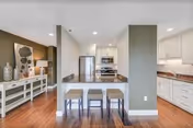 Open-plan interior showing a white kitchen with an island and three stools, stainless steel appliances, hardwood floors and a decorated console table in the adjacent living area.