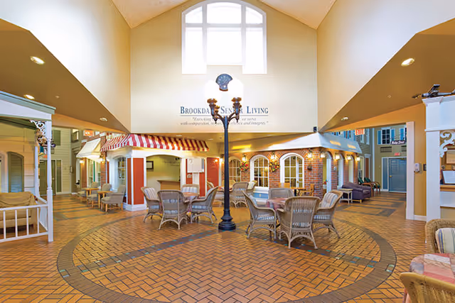 A spacious indoor common area at Brookdale Salem featuring wicker chairs and tables arranged on a patterned brick floor. The area is designed to resemble a small village street with storefront facades, awnings, and a central black street lamp. Large windows near the ceiling allow natural light to brighten the space.