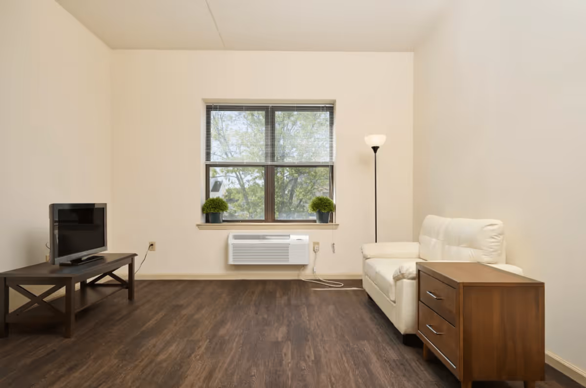 A simple living room with a white couch, a wooden side table with drawers, a floor lamp, a window with blinds and two small potted plants on the windowsill, a wall-mounted air conditioning unit below the window, and a wooden TV stand with a flat-screen television on it. The room has light-colored walls and dark wood flooring.