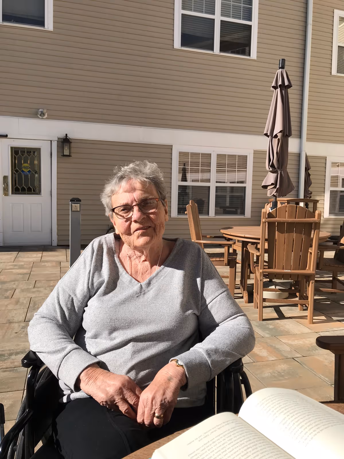 An elderly woman with glasses sitting in a wheelchair outside on a patio area of a residential building, smiling gently at the camera. Behind her are wooden outdoor chairs and tables with closed umbrellas, and windows of the building are visible.