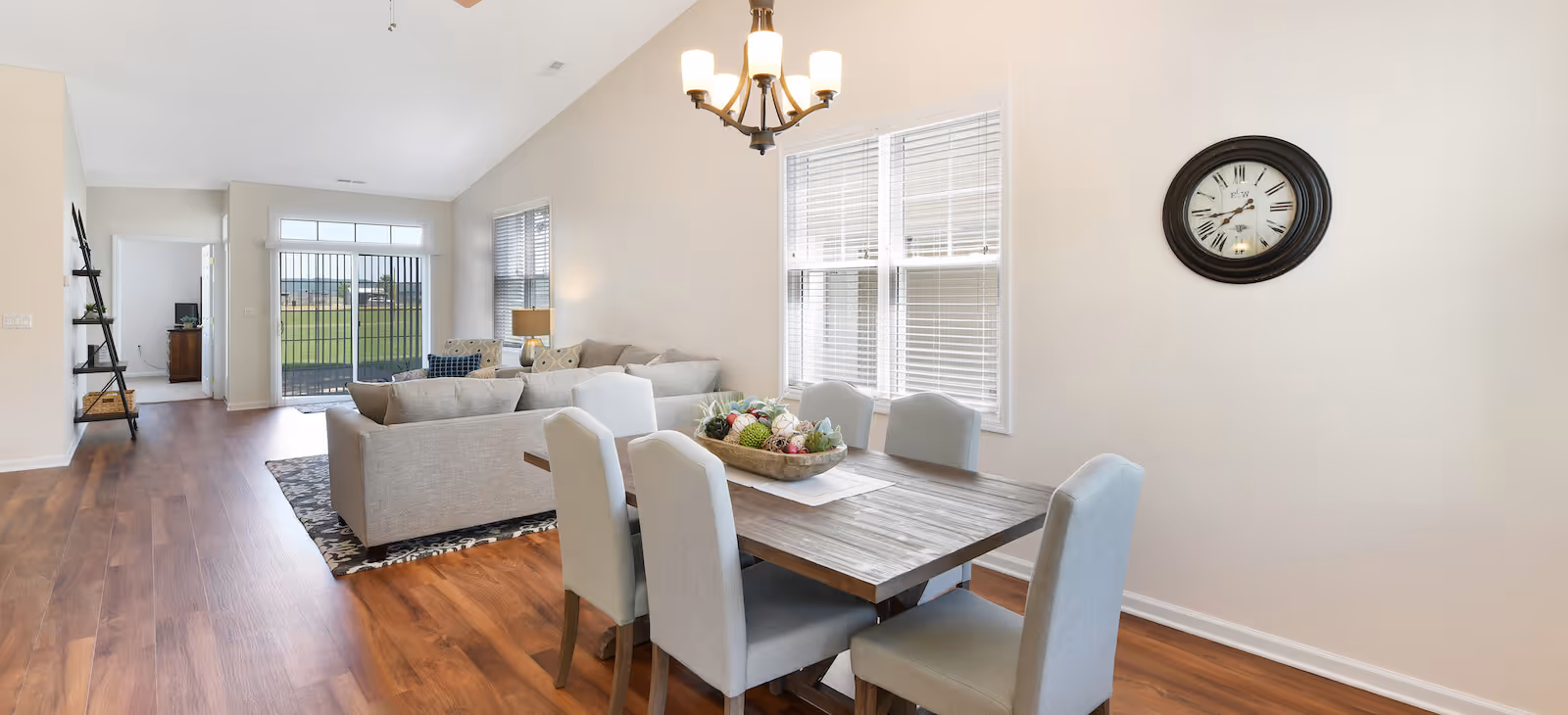 A bright and spacious interior featuring a dining area with a wooden table and six upholstered chairs. A decorative centerpiece is placed on the table. Adjacent to the dining area is a living room with a large sectional sofa, a floor lamp, and a window with blinds. The room has wooden flooring, a wall clock, and a chandelier hanging from the ceiling. Large sliding glass doors lead to an outdoor area with a grassy view.