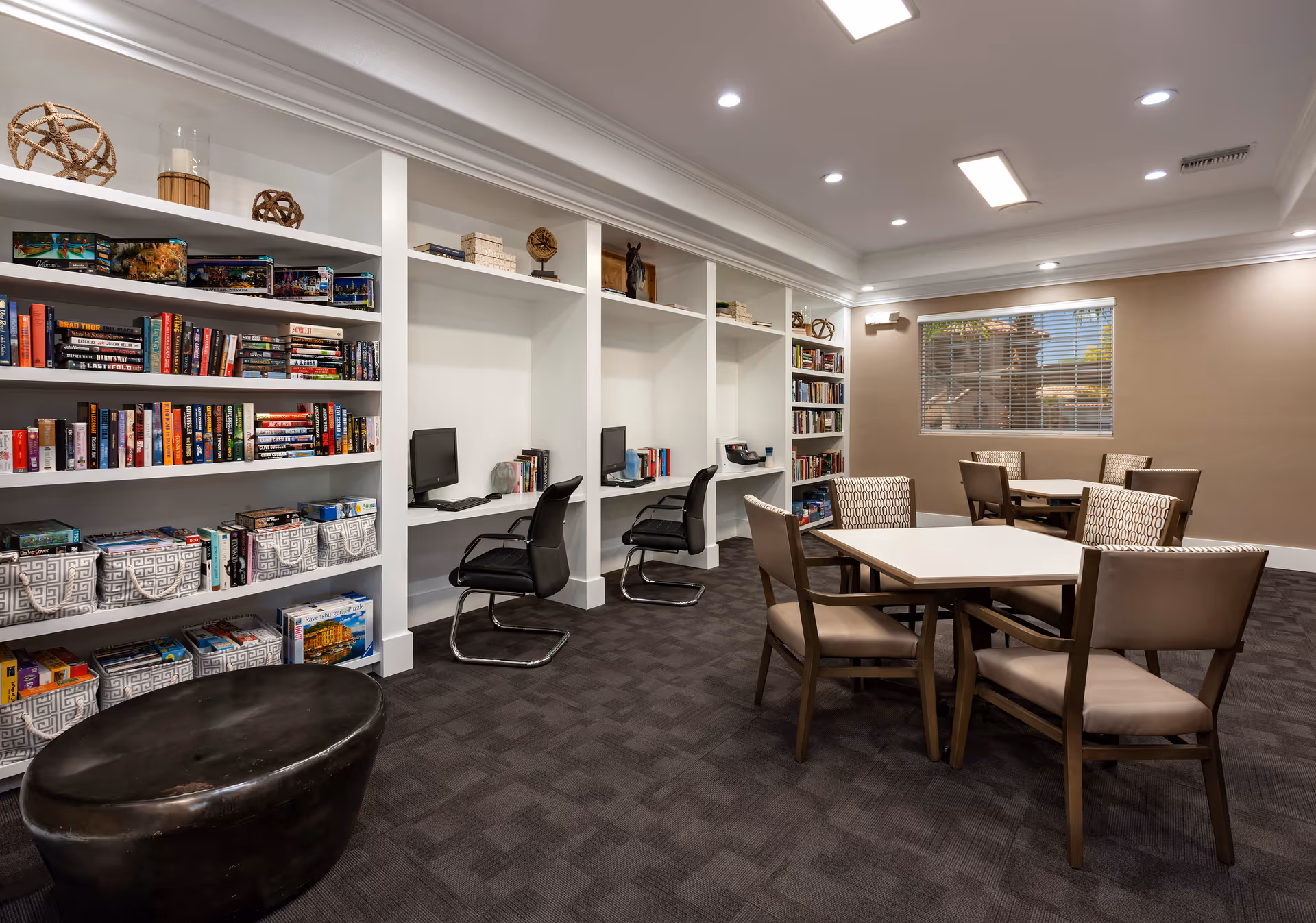 A well-lit room with bookshelves filled with books and board games, two computer desks with black chairs, and two tables surrounded by beige cushioned chairs. A window with blinds is visible on the far wall.