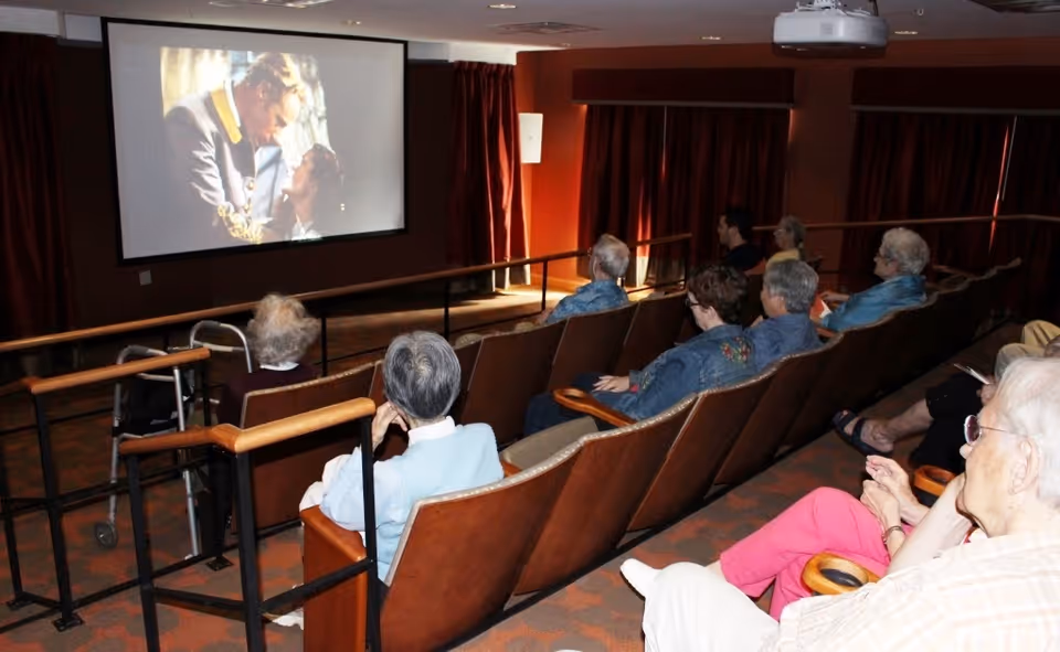 A group of elderly people seated in a small theater room watching a movie projected on a large screen. The room has brown walls, red curtains, and rows of brown cushioned seats with wooden armrests. A walker is visible next to one of the seats.