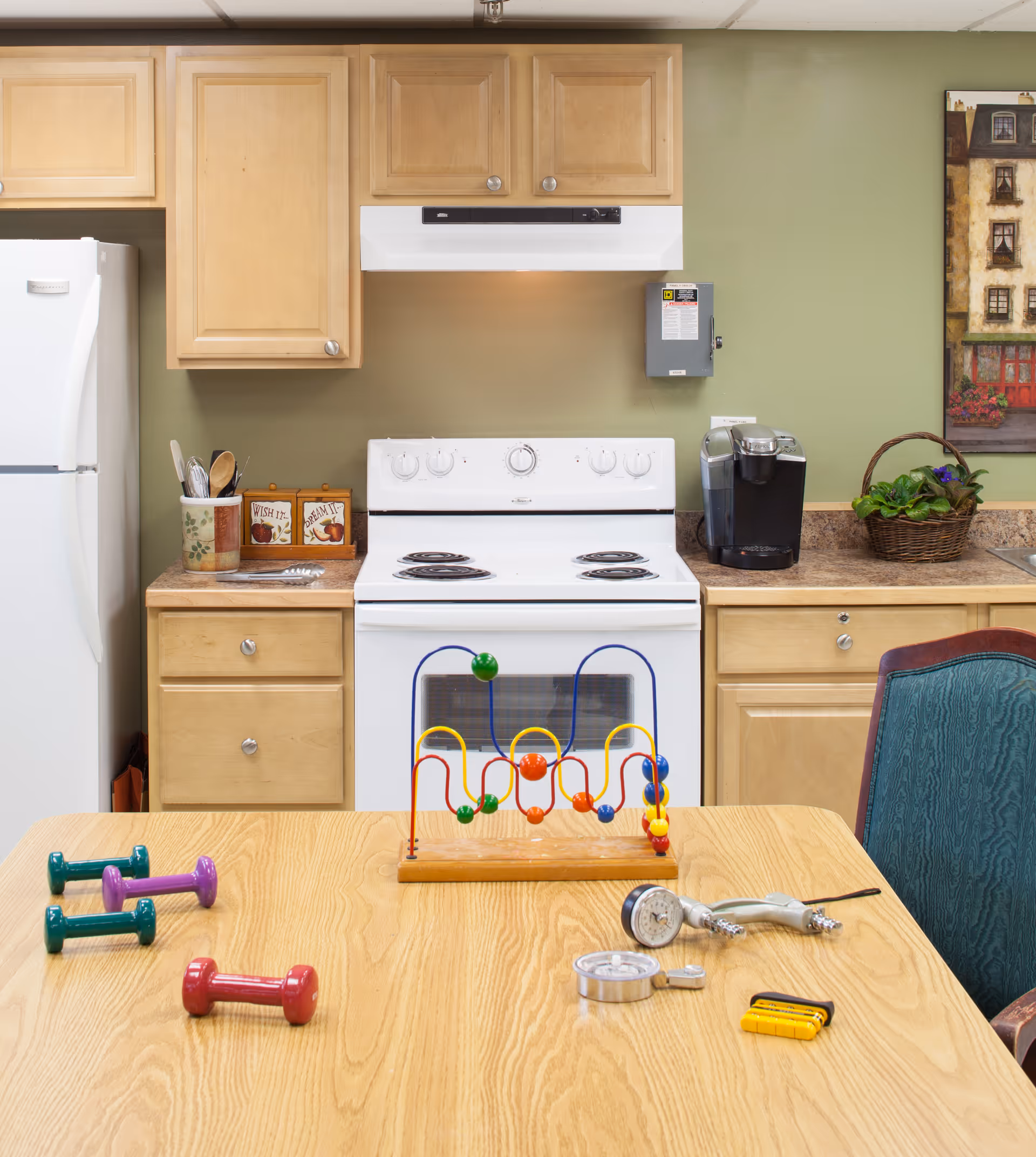 A kitchen area with light wood cabinets, a white refrigerator, and a white electric stove with an overhead vent. On the countertop to the right of the stove is a coffee maker and a basket with a plant. In the foreground, a wooden table holds colorful small dumbbells, a bead maze toy, and some medical or exercise instruments. A green upholstered chair is partially visible on the right side.