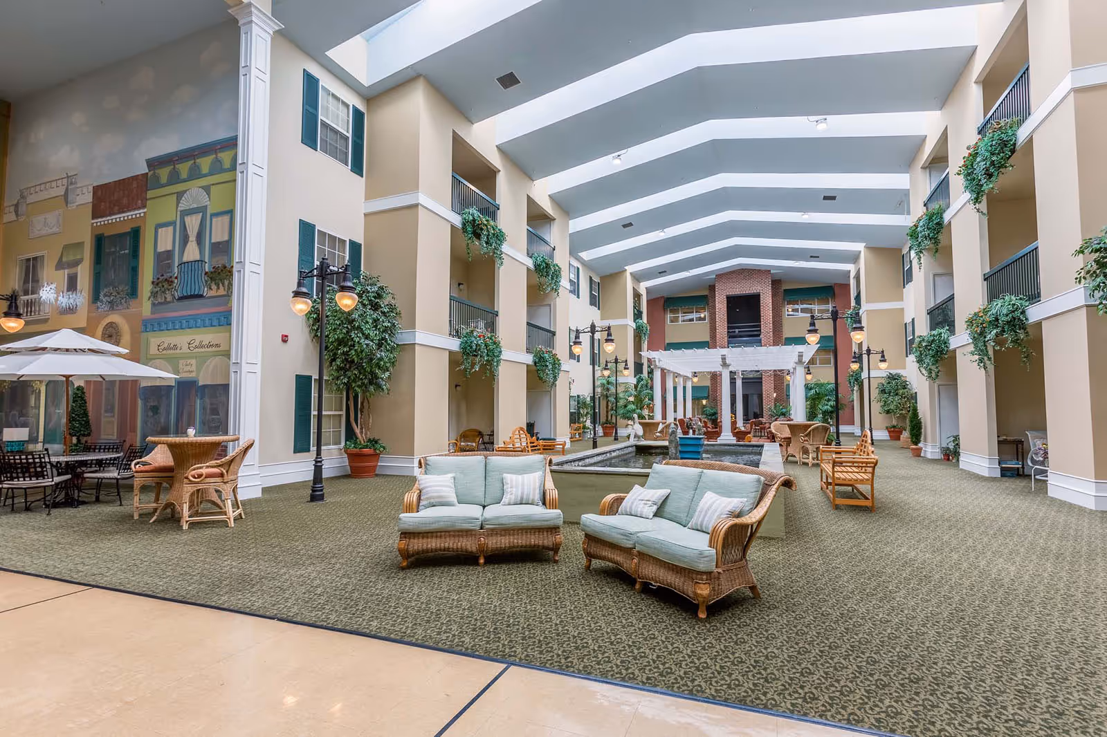 Indoor common area with high vaulted ceilings and large skylights. The space features wicker sofas with light blue cushions, several potted plants, hanging greenery, and streetlamp-style lighting. There is a central water feature with a pergola and additional seating in the background. One wall has a mural depicting storefronts and windows with green shutters.