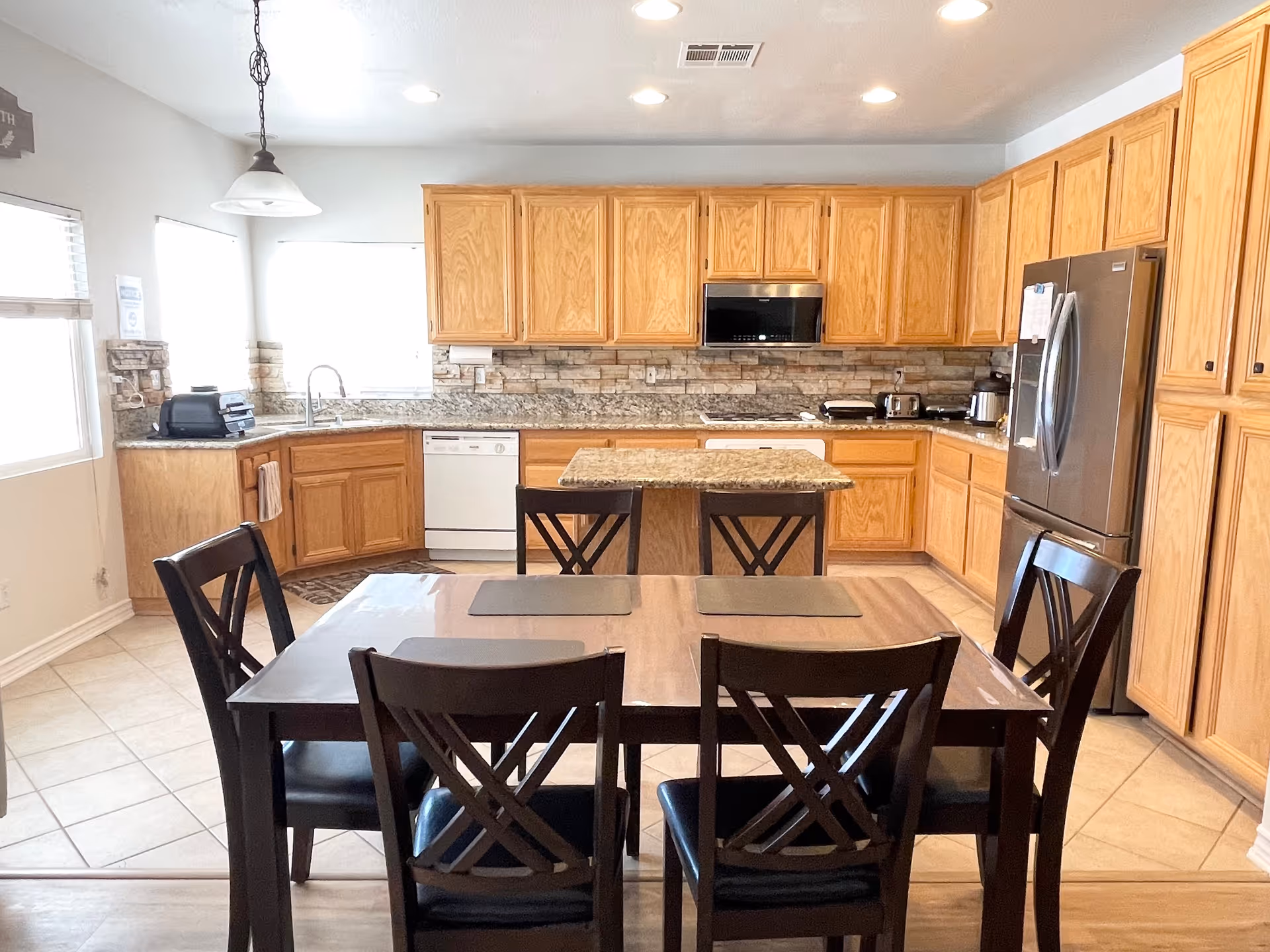 A bright kitchen with wooden cabinets, granite countertops, and stainless steel appliances including a refrigerator and microwave. There is a dining table with six dark wooden chairs in the foreground, and a kitchen island in the center. The kitchen has tiled floors and two windows letting in natural light.
