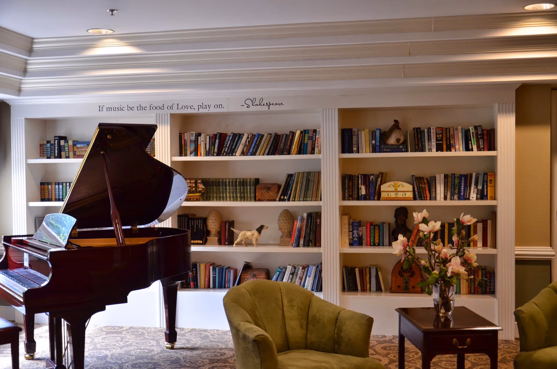 A cozy interior room featuring a grand piano on the left, a built-in bookshelf filled with books and decorative items in the background, and two green upholstered armchairs with a small wooden table holding a vase of flowers in the foreground. Above the bookshelf is a quote by Shakespeare that reads, 'If music be the food of Love, play on.'