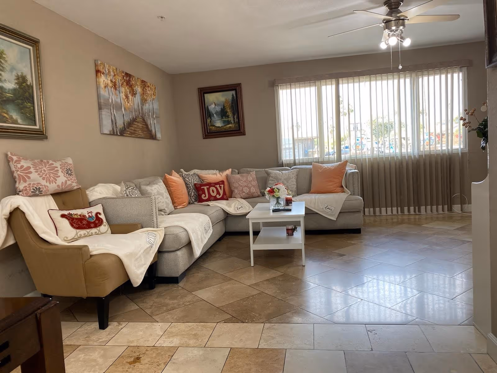 A cozy living room with a large beige sectional sofa adorned with various decorative pillows and throws. There is a beige armchair with a floral pillow and a white throw. A small white coffee table with a flower arrangement and candles sits in front of the sofa. The room has tiled flooring and three framed paintings on the walls. A ceiling fan with lights hangs from the ceiling, and large windows with vertical blinds allow natural light to fill the space.