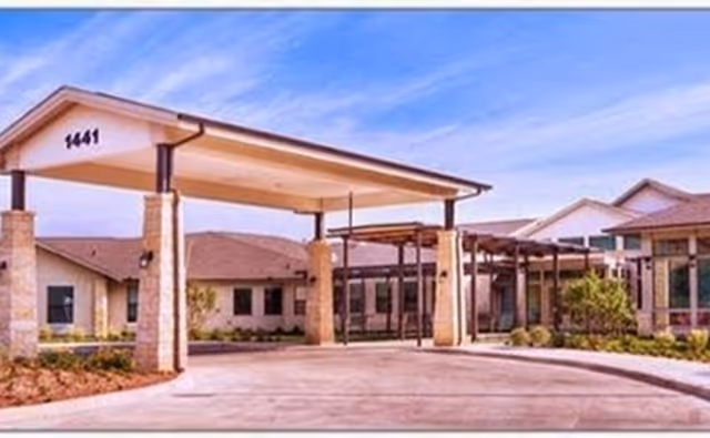Exterior view of a senior living facility entrance with a covered drop-off area supported by stone pillars, a driveway, and a clear blue sky.