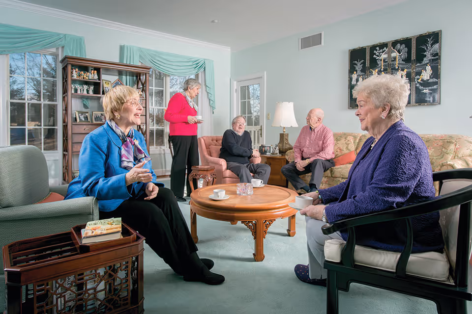A group of five elderly people sitting and standing in a well-lit living room, engaged in conversation. The room has large windows with green curtains, a wooden coffee table with cups, comfortable chairs, a sofa, and decorative items including a lamp and wall art.