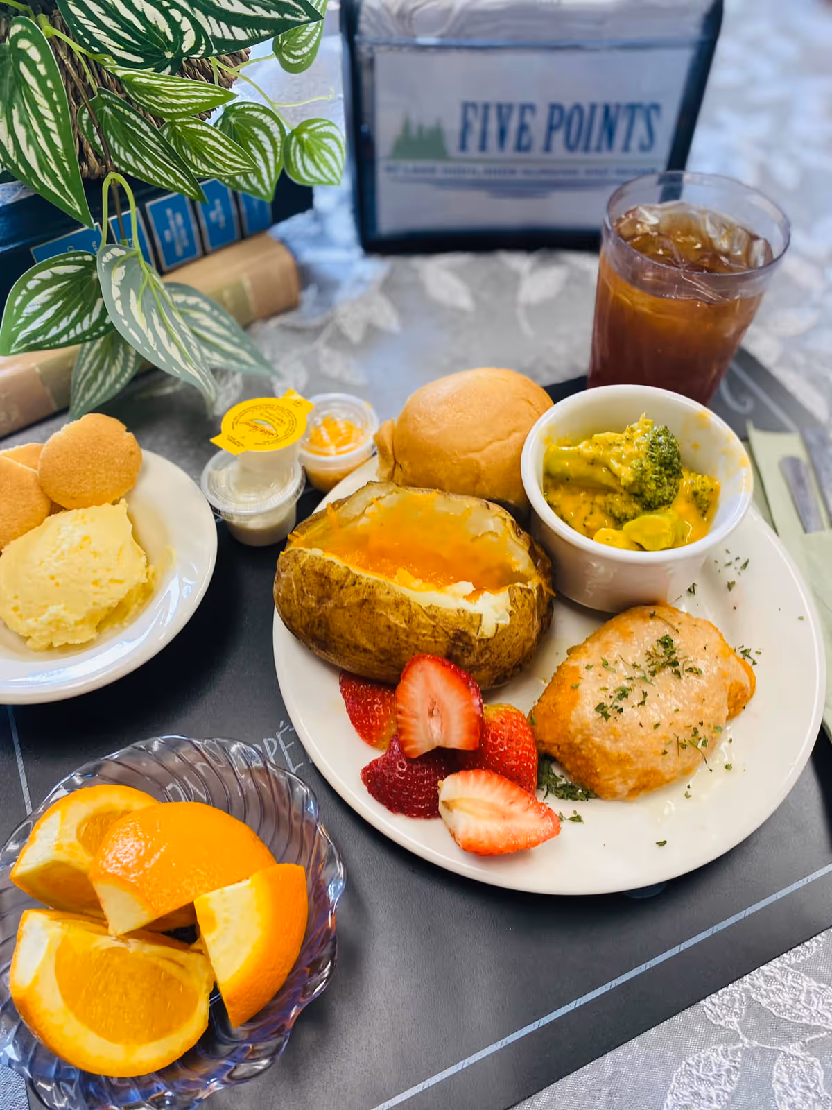 A meal served on a tray with a baked potato topped with cheese, a bread roll, a small bowl of broccoli with cheese sauce, a piece of cooked chicken garnished with herbs, and sliced strawberries on a white plate. Next to the plate is a small bowl with orange slices, a dish with mashed potatoes and small round crackers, and a glass of iced tea. In the background, there is a plant and a napkin holder with the text 'FIVE POINTS'.