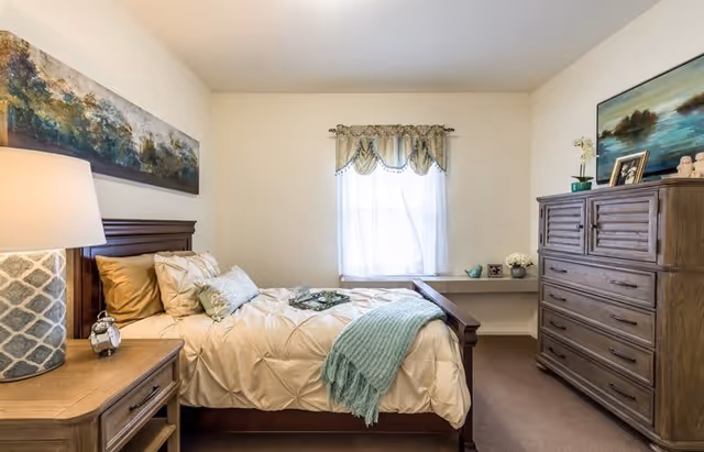 A cozy bedroom with a wooden bed frame and beige bedding accented with a light blue throw blanket and pillows. There is a wooden nightstand with a patterned lamp and an alarm clock. A large wooden dresser with decorative items and a framed picture is on the right side. A window with sheer curtains and a valance lets in natural light. The walls are light-colored, and there is a landscape painting above the bed.