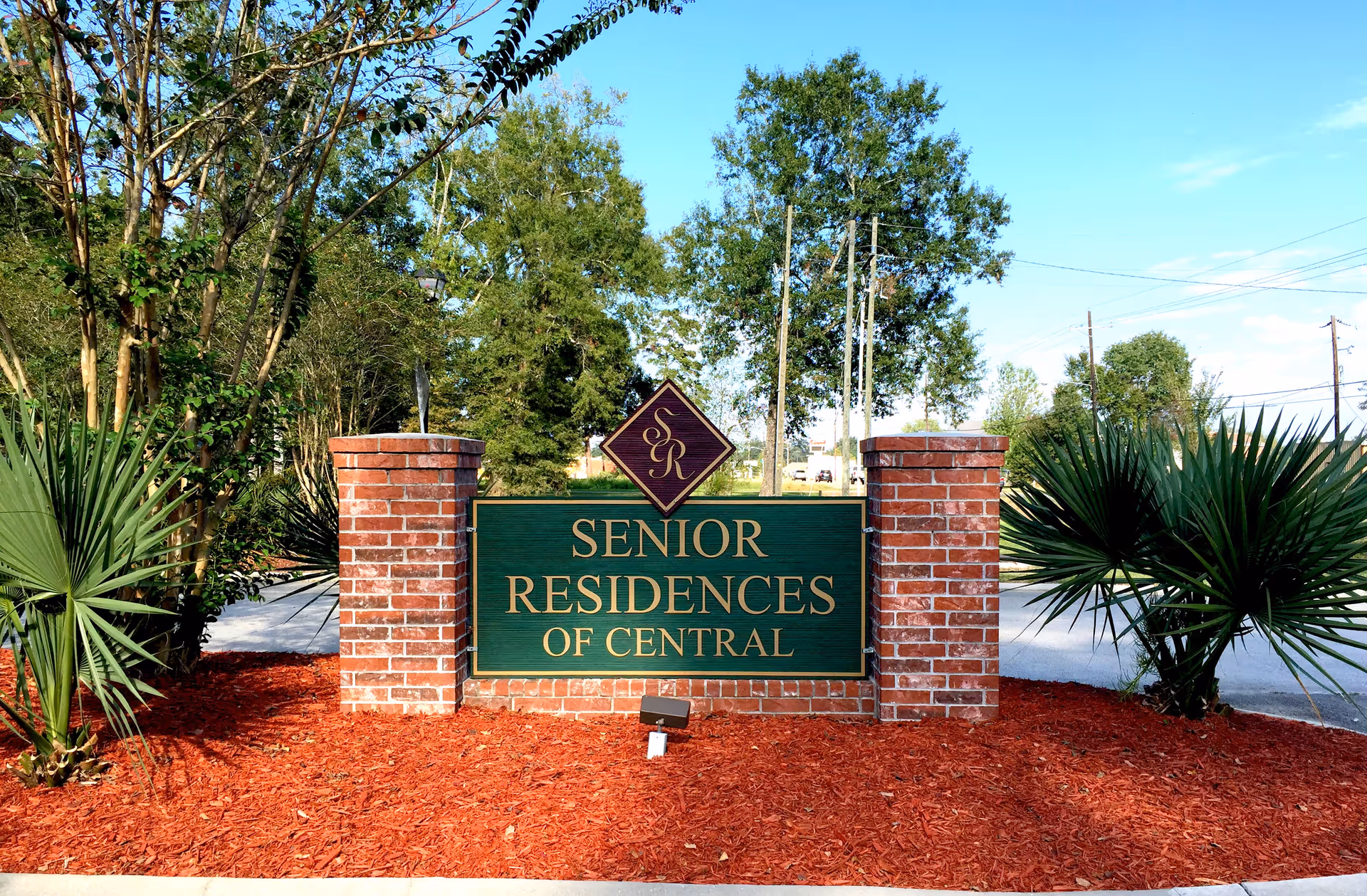 Entrance sign for Senior Residences of Central made of green and gold lettering mounted on a brick base, surrounded by landscaped plants and mulch with trees and a clear blue sky in the background.