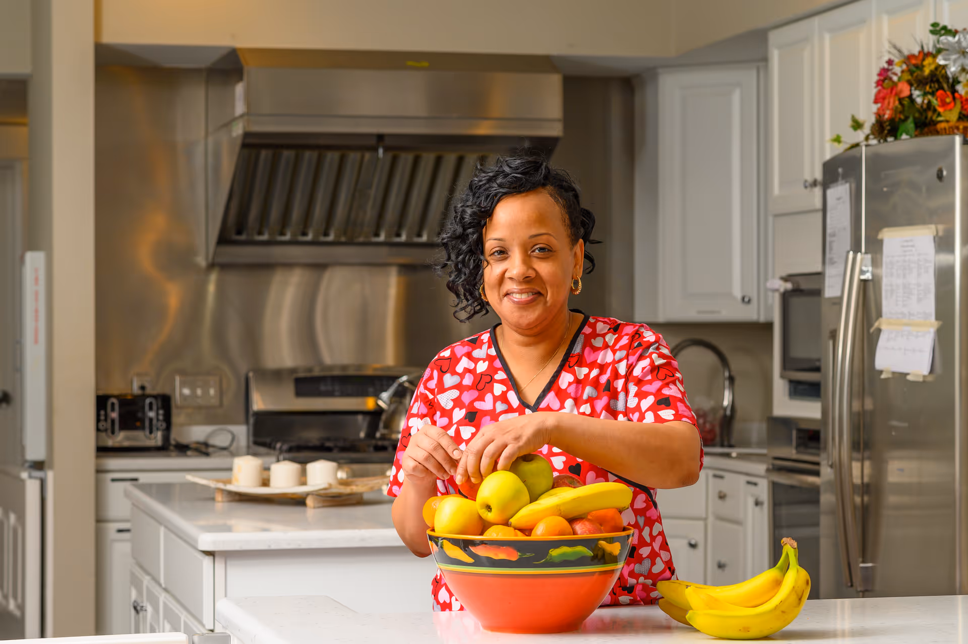 A woman wearing a red shirt with white hearts is standing in a kitchen, smiling while arranging fruit in a large bowl on a white countertop. The kitchen has white cabinets, a stainless steel refrigerator, and a stove with a stainless steel hood.