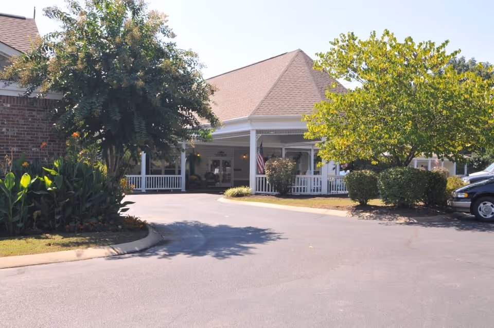 Exterior view of Charter Senior Living of Jackson showing the front entrance with a covered porch, surrounded by trees, bushes, and a paved driveway with a parked vehicle on the right.
