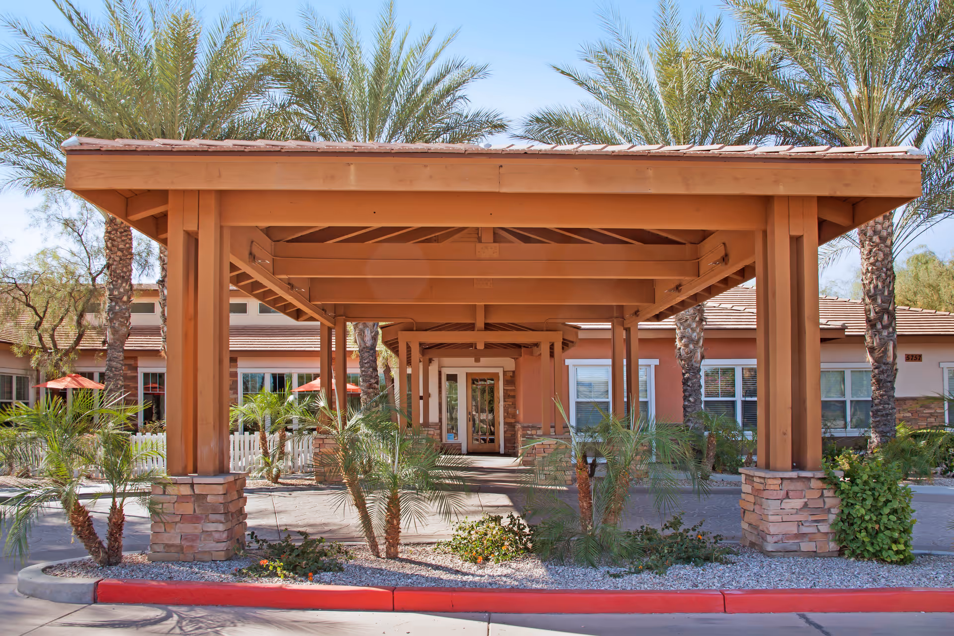 Covered wooden porte-cochère entrance with palm trees and a single-story building facade behind it.