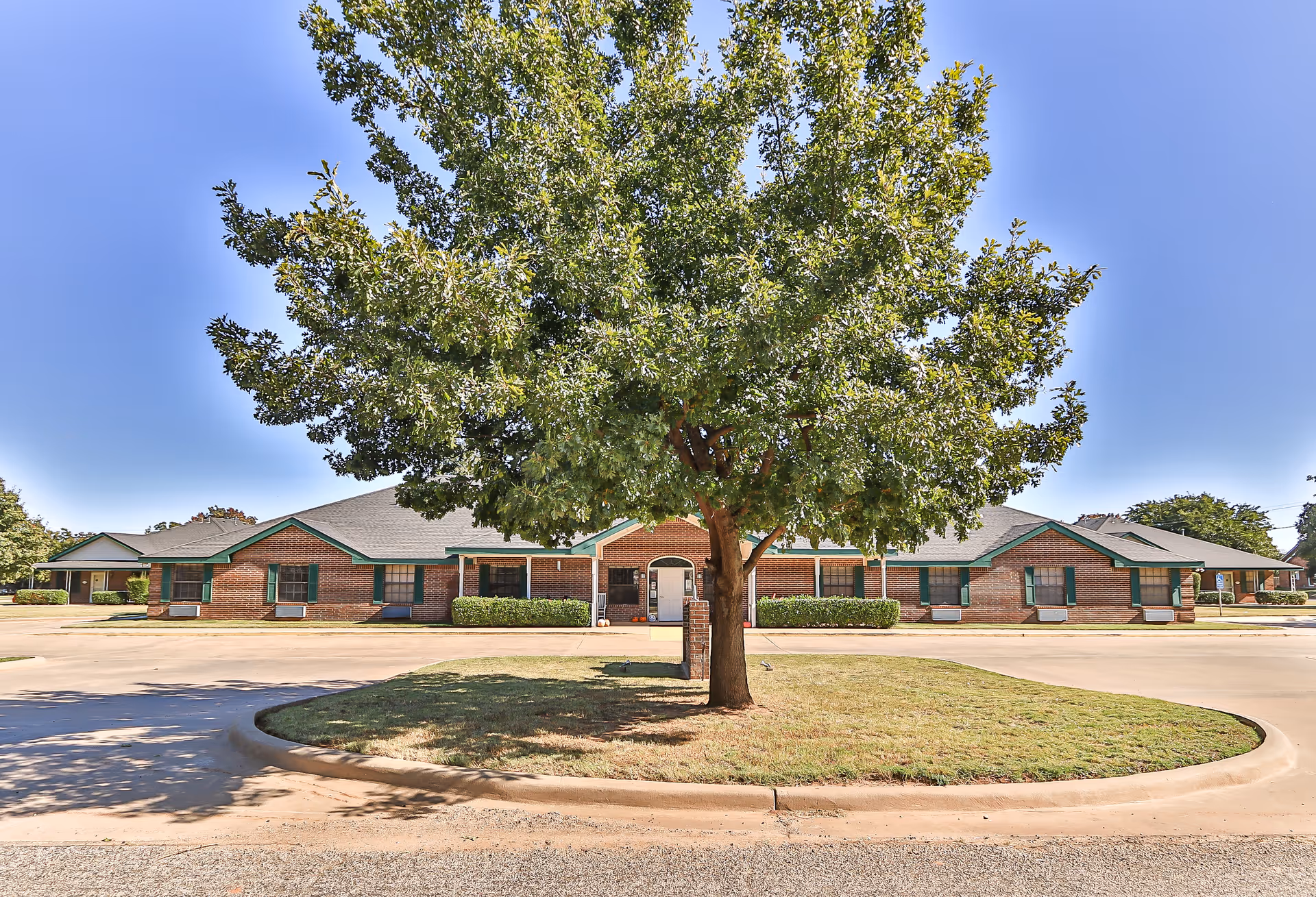 Front view of a single-story brick senior living building with green trim and a large tree centered in the foreground.