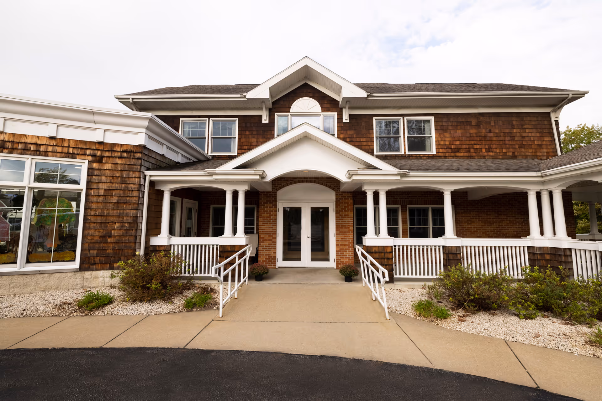 Front exterior view of a two-story senior care facility building with wooden shingles and brick walls, featuring a covered entrance with white columns and railings, and a paved walkway leading to double glass doors.