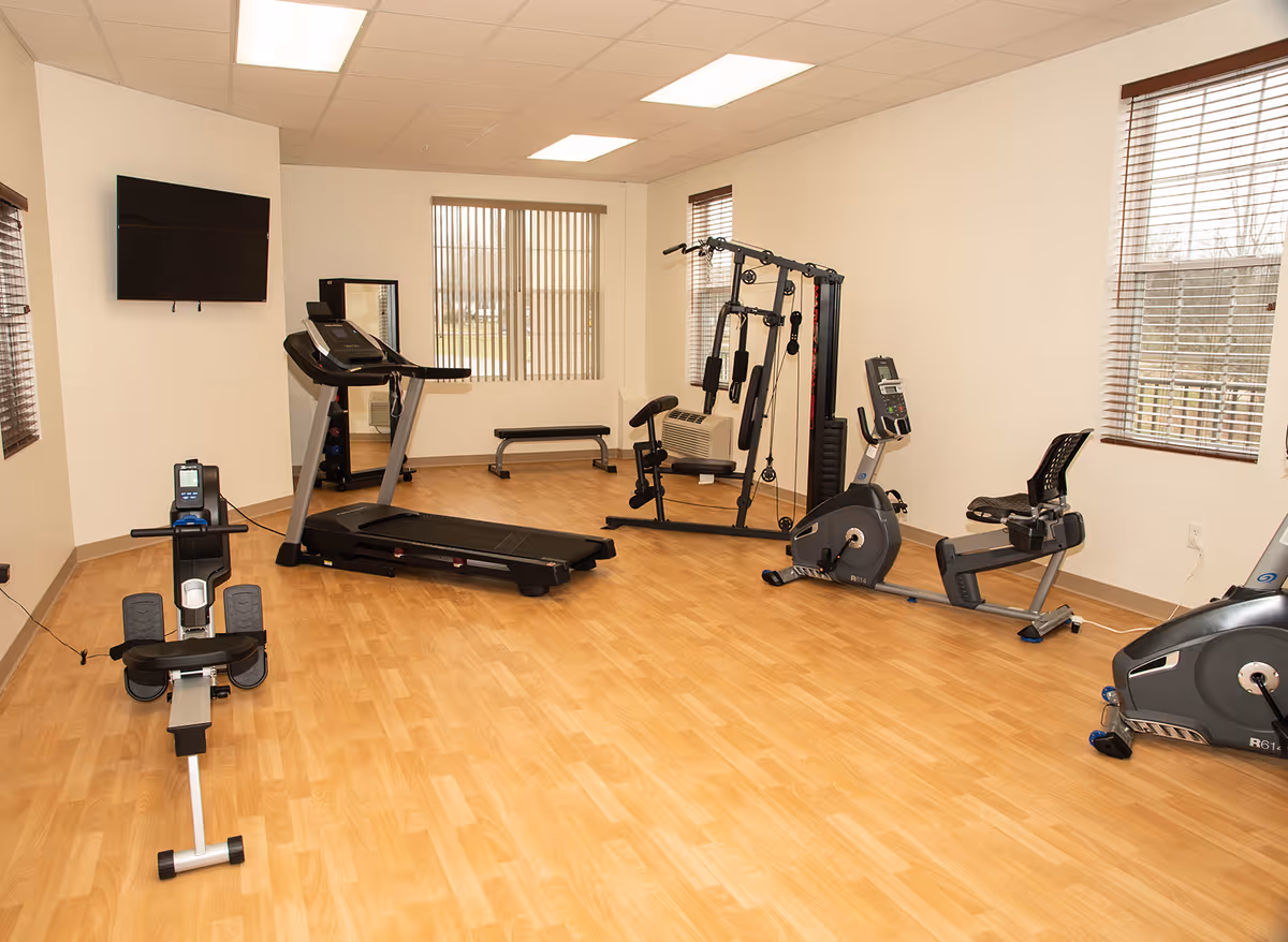 A fitness room with wooden flooring and white walls featuring various exercise equipment including a rowing machine, treadmill, multi-gym machine, stationary bike, and recumbent bike. The room has three windows with blinds and a wall-mounted flat screen TV.