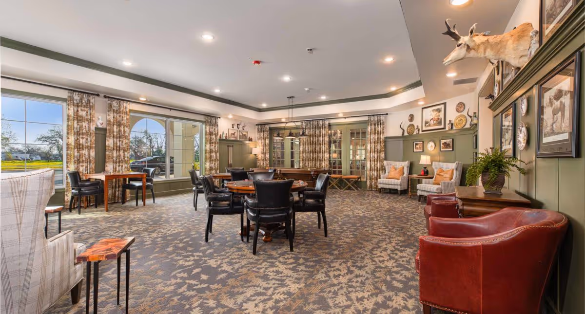 Spacious senior living communal dining and lounge area with tables, chairs, armchairs, patterned carpet and large windows.