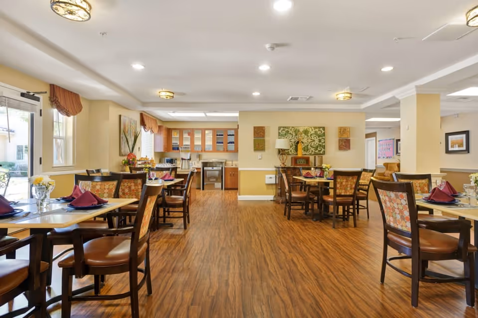 A bright and spacious dining room in a senior living facility with wooden floors, multiple tables set with plates, glasses, and burgundy napkins. The room features large windows with valances, warm yellow walls, and decorative artwork. The back of the room has a kitchen area with wooden cabinets and appliances.