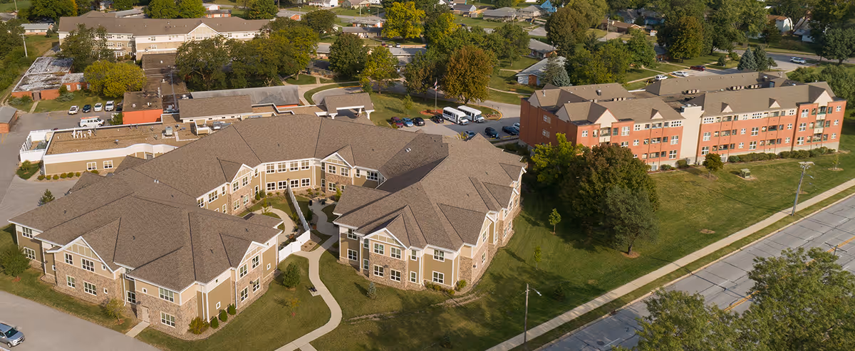 Aerial view of Village Assisted Living facility showing multiple connected buildings with beige and brick exteriors, surrounded by green lawns, trees, parking areas, and nearby roads.