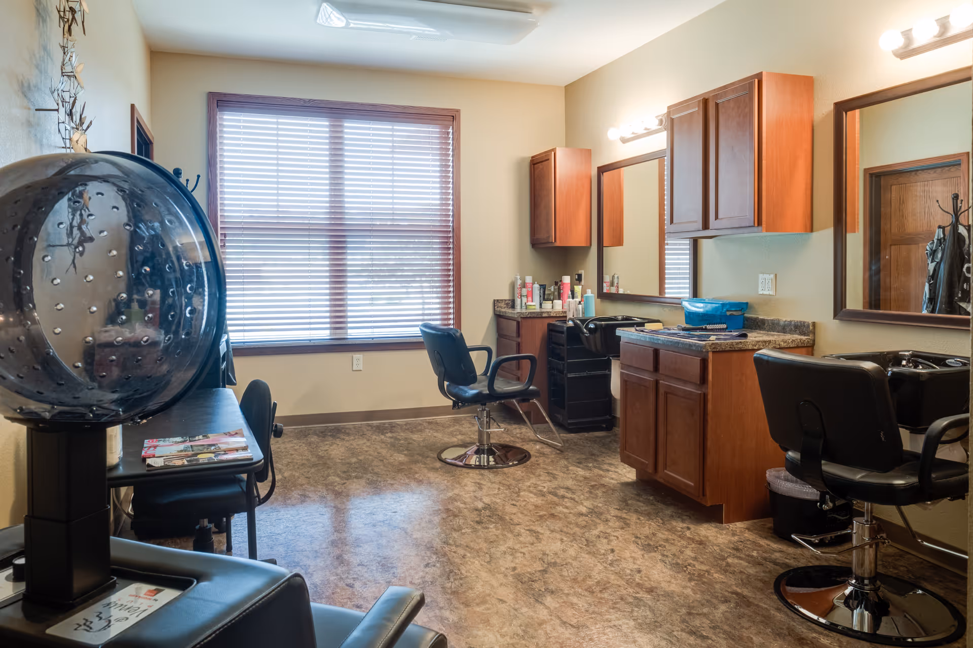 Interior of a senior living facility hair salon with two black salon chairs, hair washing sinks, wooden cabinets, a large window with blinds, and a hair dryer chair in the foreground.