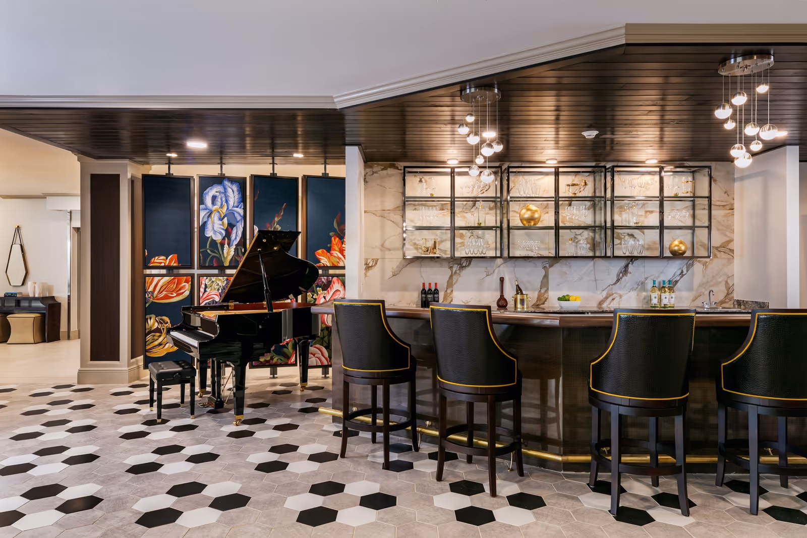 Interior view of a stylish lounge area featuring a black grand piano with a matching bench, a decorative floral panel behind it, and a bar counter with four black and gold bar stools. The bar has glass shelves with glassware and decorative items, and modern pendant lights hang from the dark wooden ceiling. The floor has a hexagonal tile pattern in black, white, and gray.