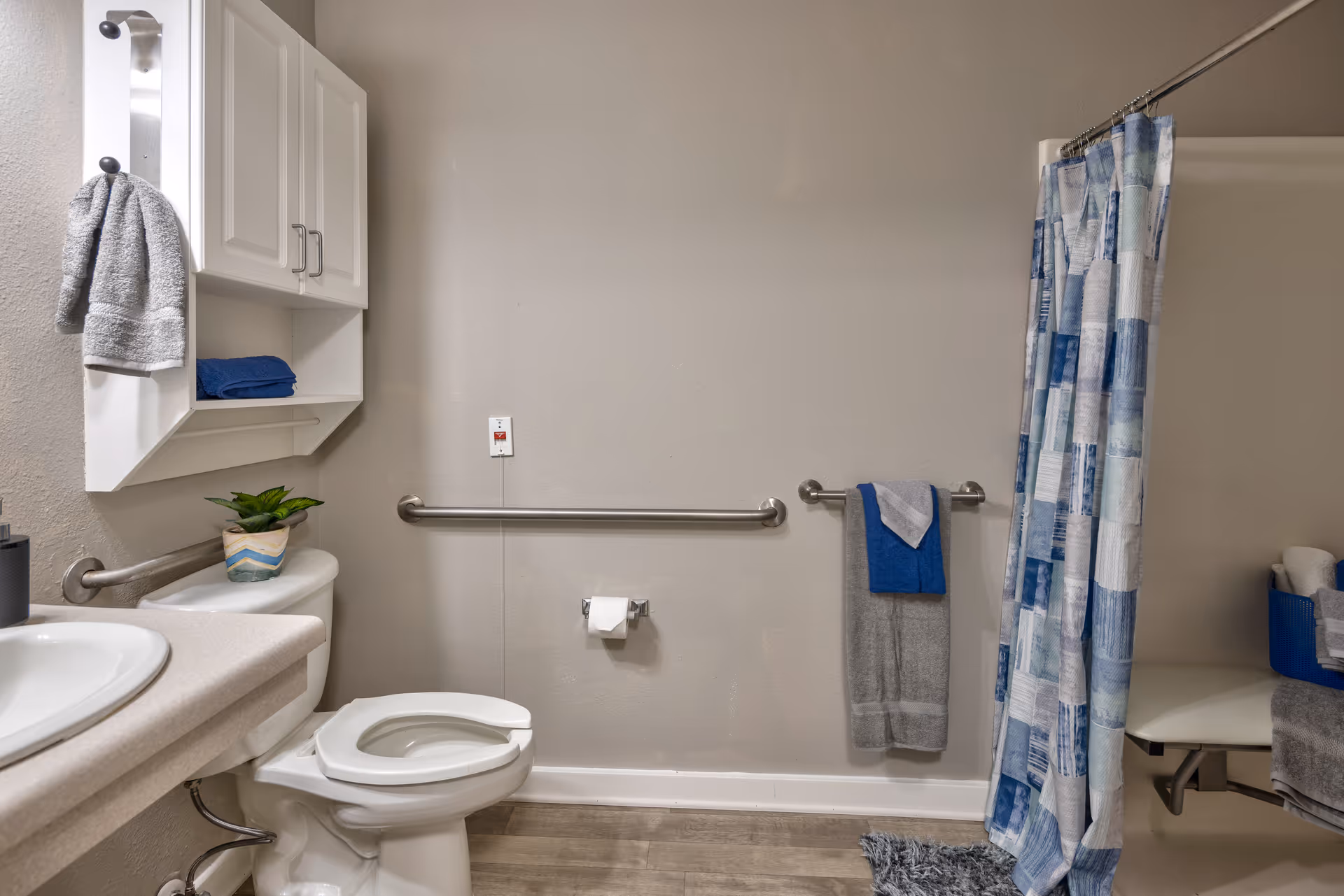 A clean and accessible bathroom featuring a white toilet with grab bars on the walls, a sink with a countertop, a white wall cabinet with towels, a shower area with a blue and white patterned curtain, and a small plant on the toilet tank.