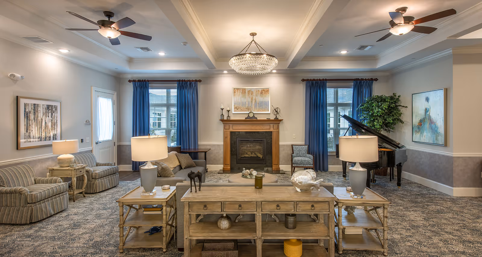 A spacious and elegantly decorated living room with a fireplace centered on the far wall. The room features two ceiling fans with lights, blue curtains on windows flanking the fireplace, a grand piano in the right corner, and various seating options including striped armchairs and a gray sofa. There are two wooden side tables with lamps and decorative items on a wooden console table in the foreground. The walls are adorned with framed artwork and the room has a patterned carpet.