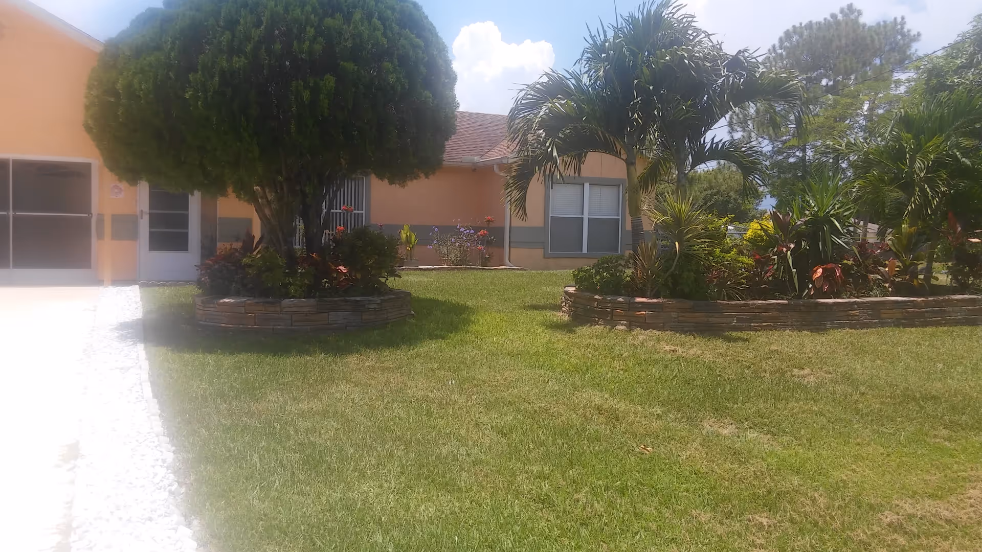 Front yard and facade of a single-story peach-colored building with a manicured lawn, circular raised flower beds and palm and trimmed trees.