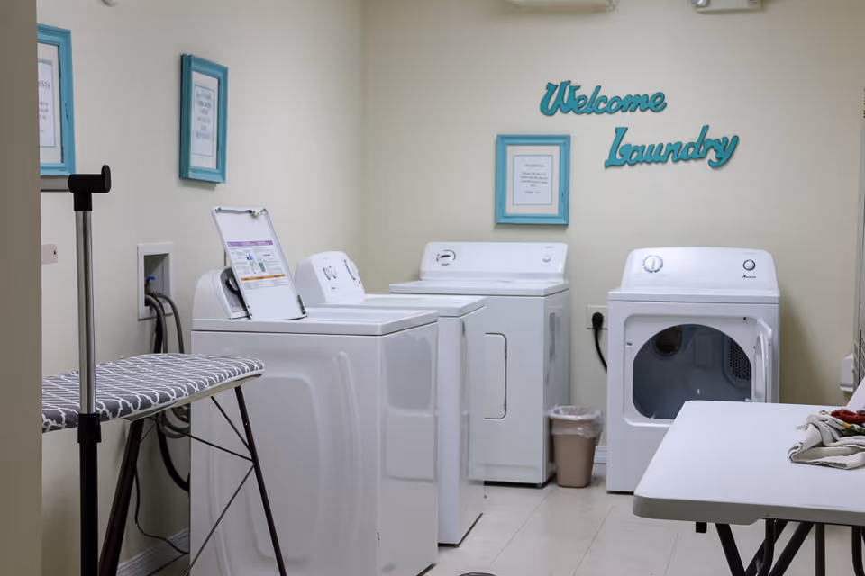 A clean laundry room with washers and a dryer, an ironing board, folding table and a wall sign that says 'Welcome Laundry'.