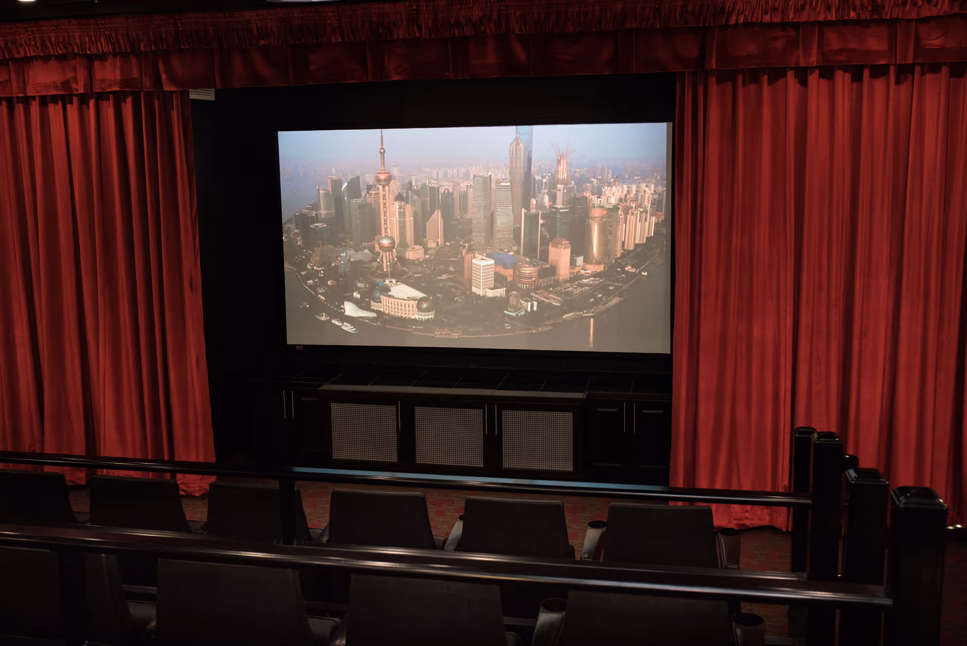 Interior view of a small theater room with red curtains on the sides, a large screen displaying a cityscape, and rows of black seats facing the screen.