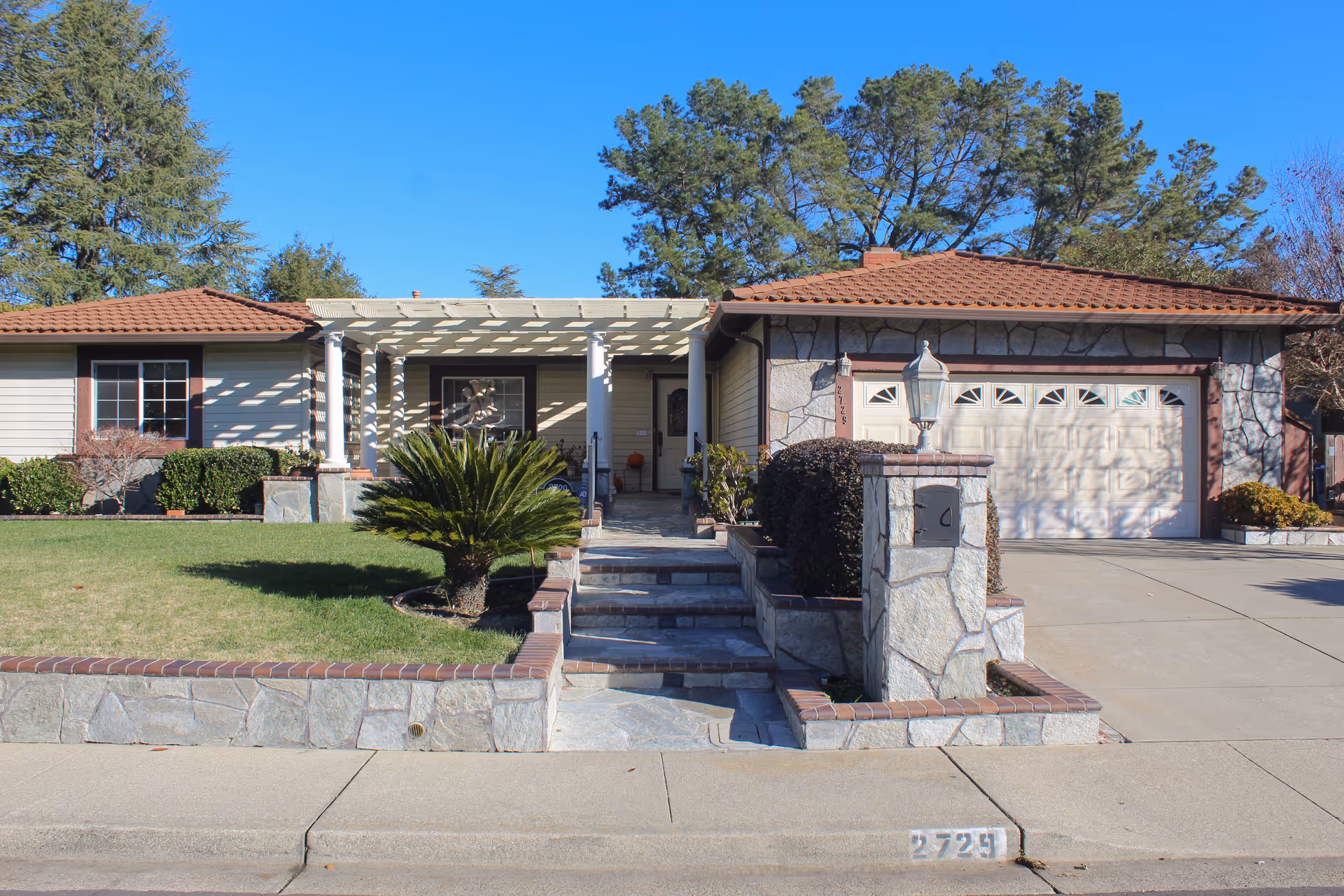 Front exterior view of a single-story house with a tiled roof, stone and brick accents, a small front lawn with a palm-like plant, a stone walkway with steps leading to the front door, and a two-car garage on the right side.