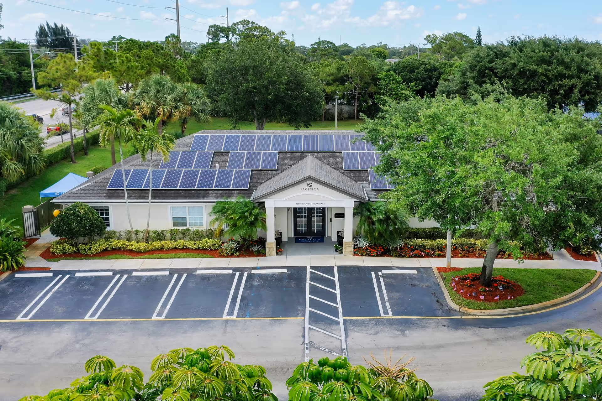 Aerial view of a single-story building with solar panels on the roof, surrounded by lush green trees and landscaping. The building has a covered entrance with a sign that reads 'Pacifica Senior Living Palm Beach.' There is a parking lot with marked spaces in front of the building.