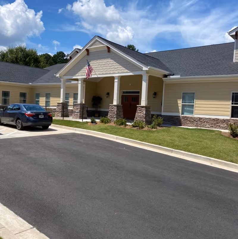 Exterior view of a single-story senior living facility building with beige siding and stone accents. The entrance features a covered porch with white columns and an American flag hanging above. A black car is parked in the parking lot in front of the building, and the sky is partly cloudy.