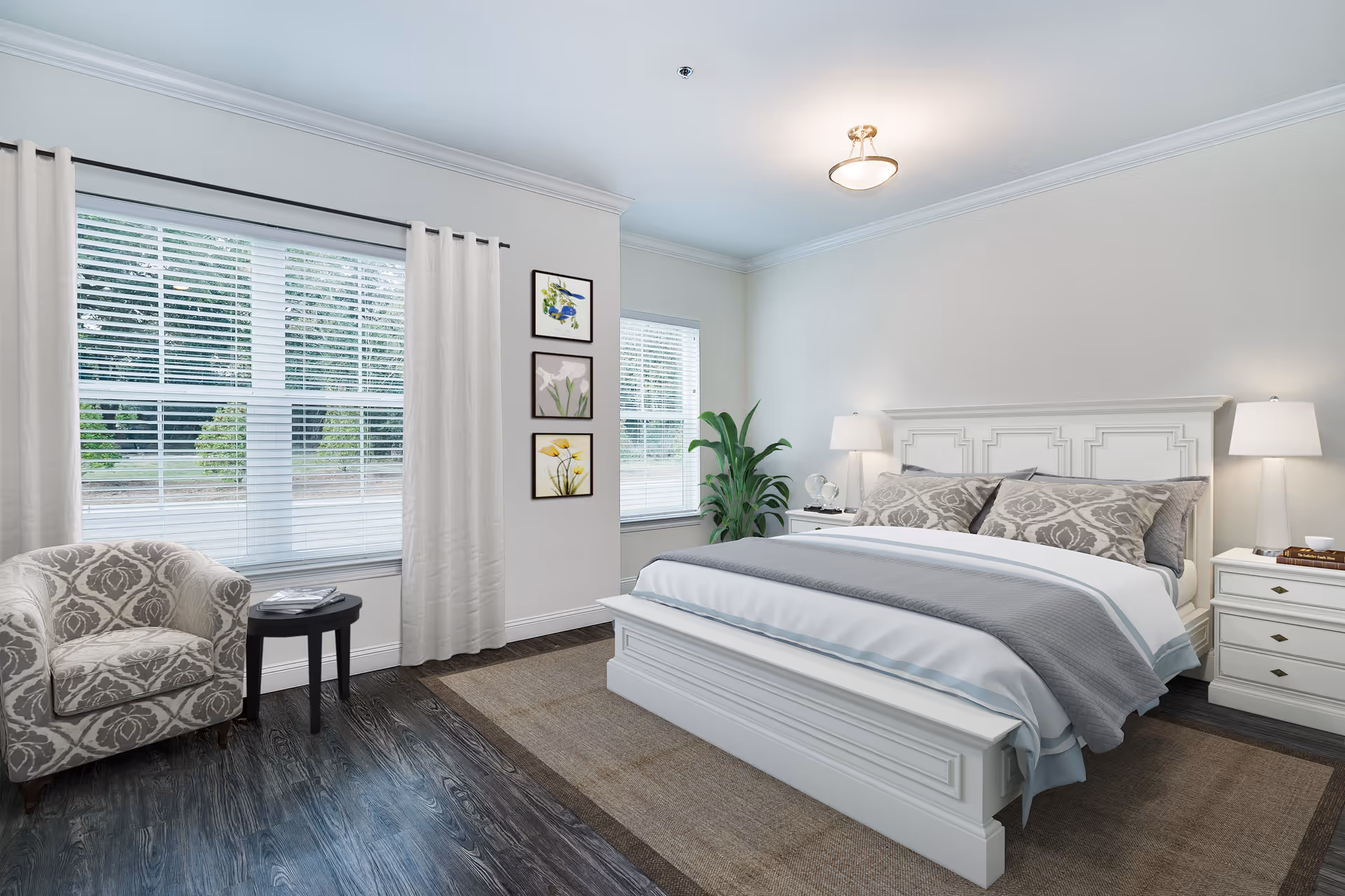 A bright and neatly arranged bedroom with a large white bed featuring gray and white bedding. There are two white nightstands with lamps on each side of the bed. A patterned armchair and a small round black table are placed near two large windows with white blinds and curtains. Three framed floral artworks hang on the wall between the windows, and a green potted plant is positioned near the bed. The room has dark wood flooring and a beige area rug under the bed.