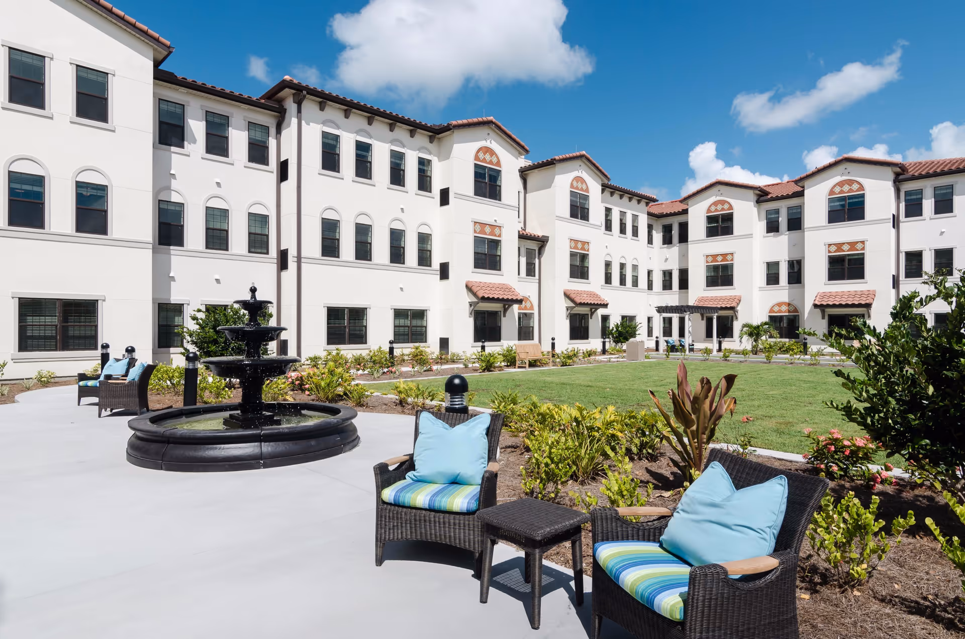 Outdoor courtyard with a central fountain, wicker chairs with cushions, and a three-story white building in the background under a blue sky.