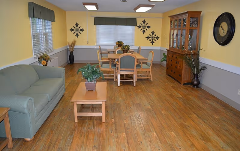 A cozy common area with wooden flooring, yellow and gray walls, a green upholstered sofa, a wooden coffee table with a potted plant, a wooden dining table with six chairs, a wooden china cabinet, wall clock, and decorative wall art.