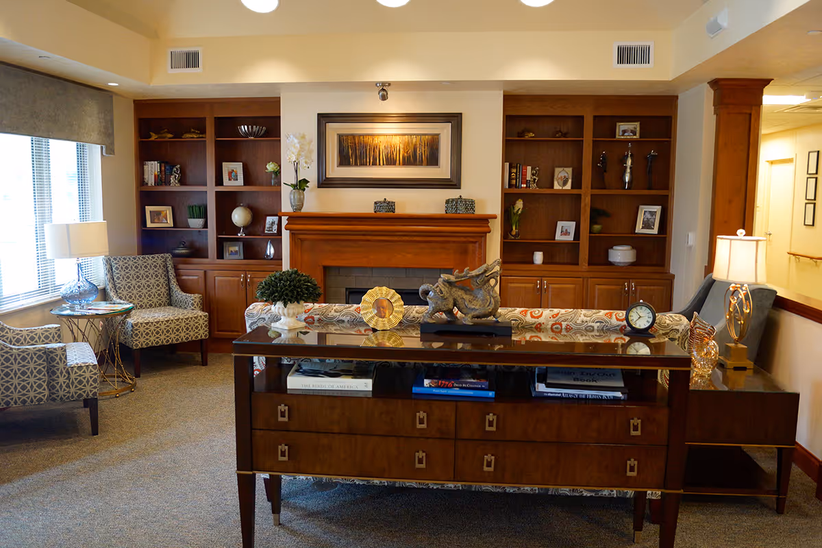 A cozy living room area with a wooden fireplace flanked by built-in bookshelves filled with books, framed photos, and decorative items. In front of the fireplace is a patterned sofa with a wooden console table behind it, displaying various decorative objects including a clock and a sculpture. Two patterned armchairs and a side table with a lamp are positioned near a window with blinds. The room has warm lighting and a carpeted floor.
