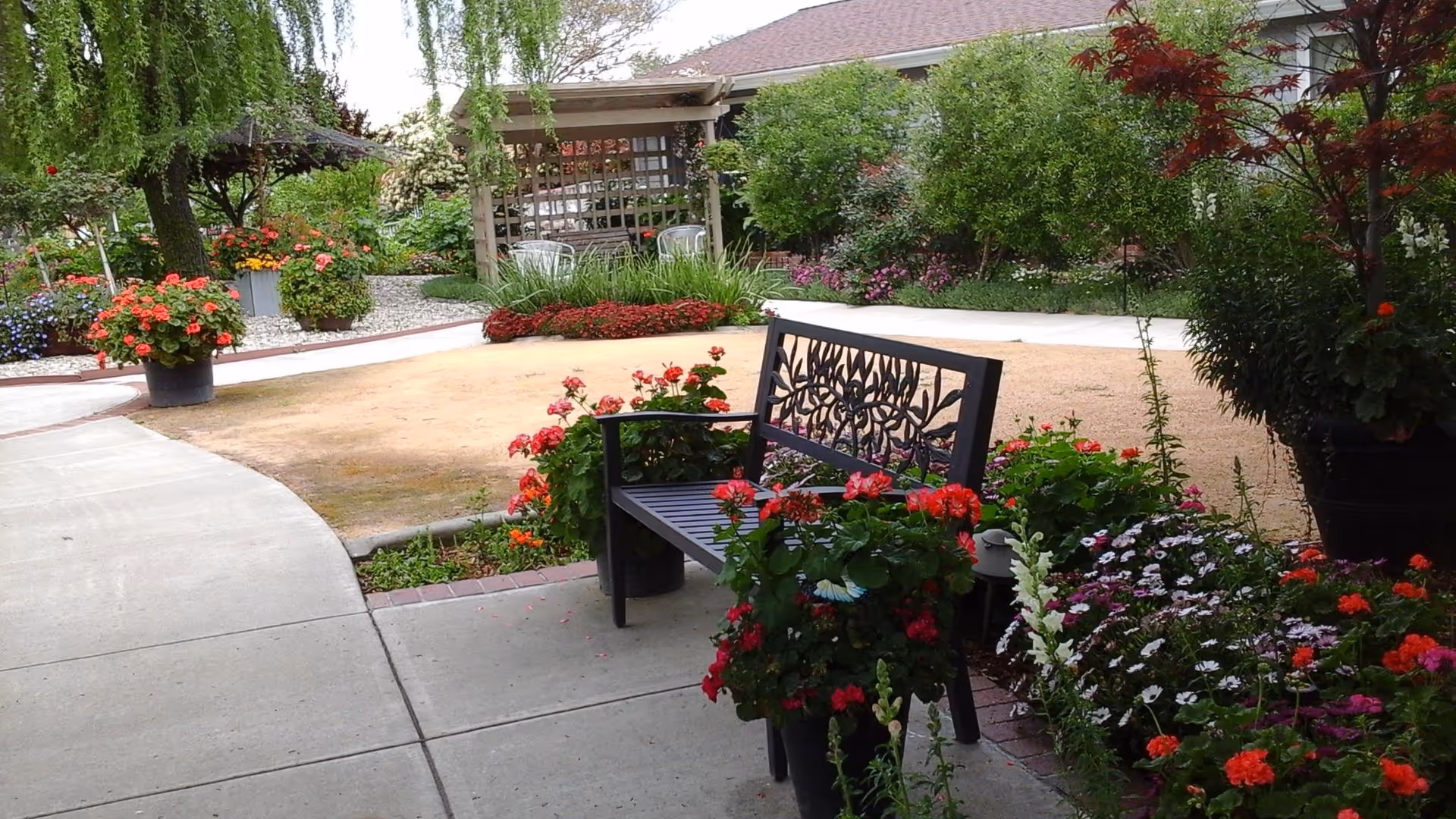 A peaceful outdoor garden area with a decorative metal bench surrounded by vibrant flowering plants in pots and garden beds. A curved concrete pathway leads through the garden, with a wooden pergola and additional seating visible in the background. The scene is lush with greenery and colorful flowers.