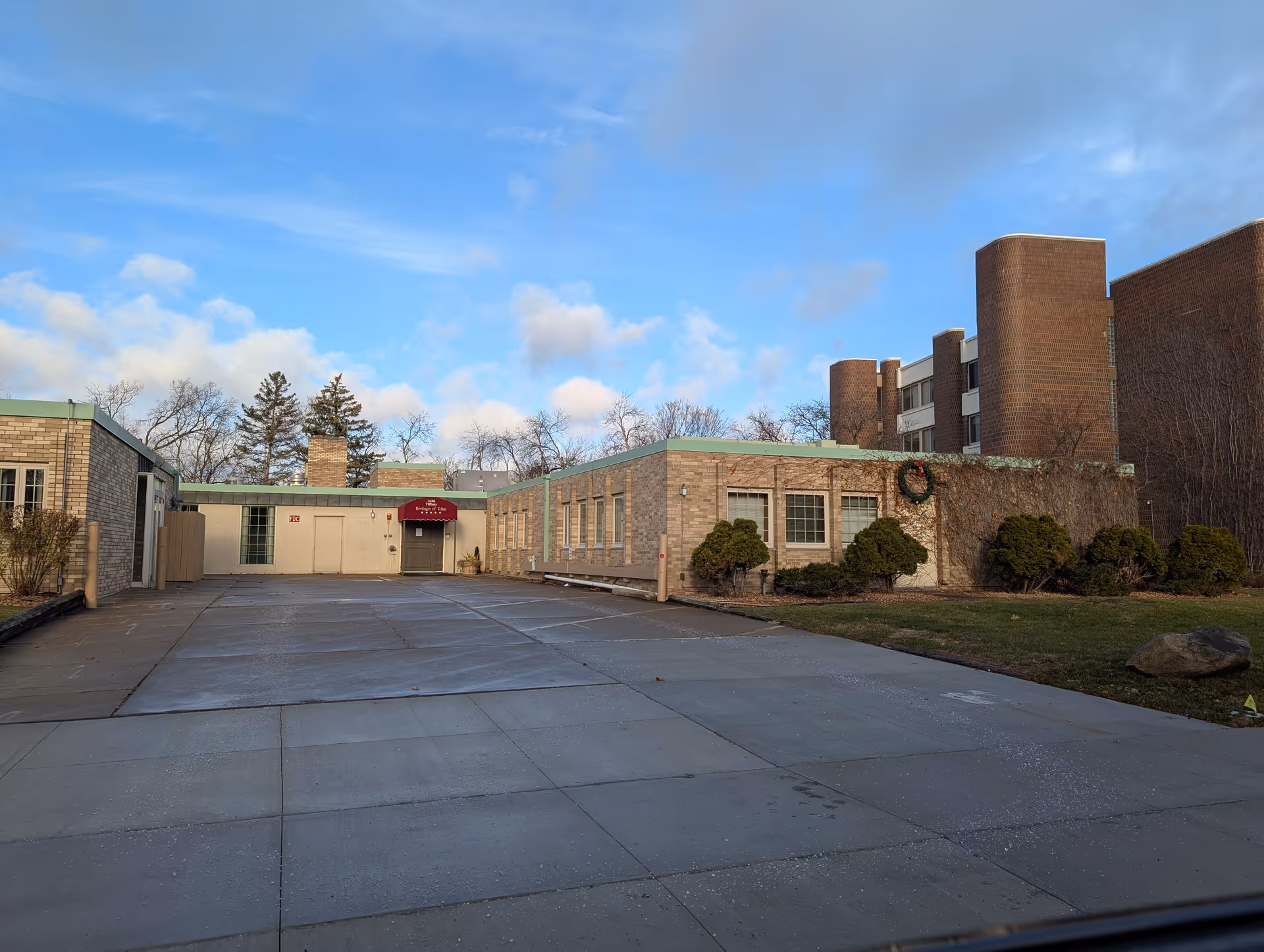 Exterior view of a senior living facility building with a paved driveway, brick walls, several windows, and a red awning above the entrance door. There are some bushes and a lawn area on the right side, with a clear blue sky and some clouds in the background.