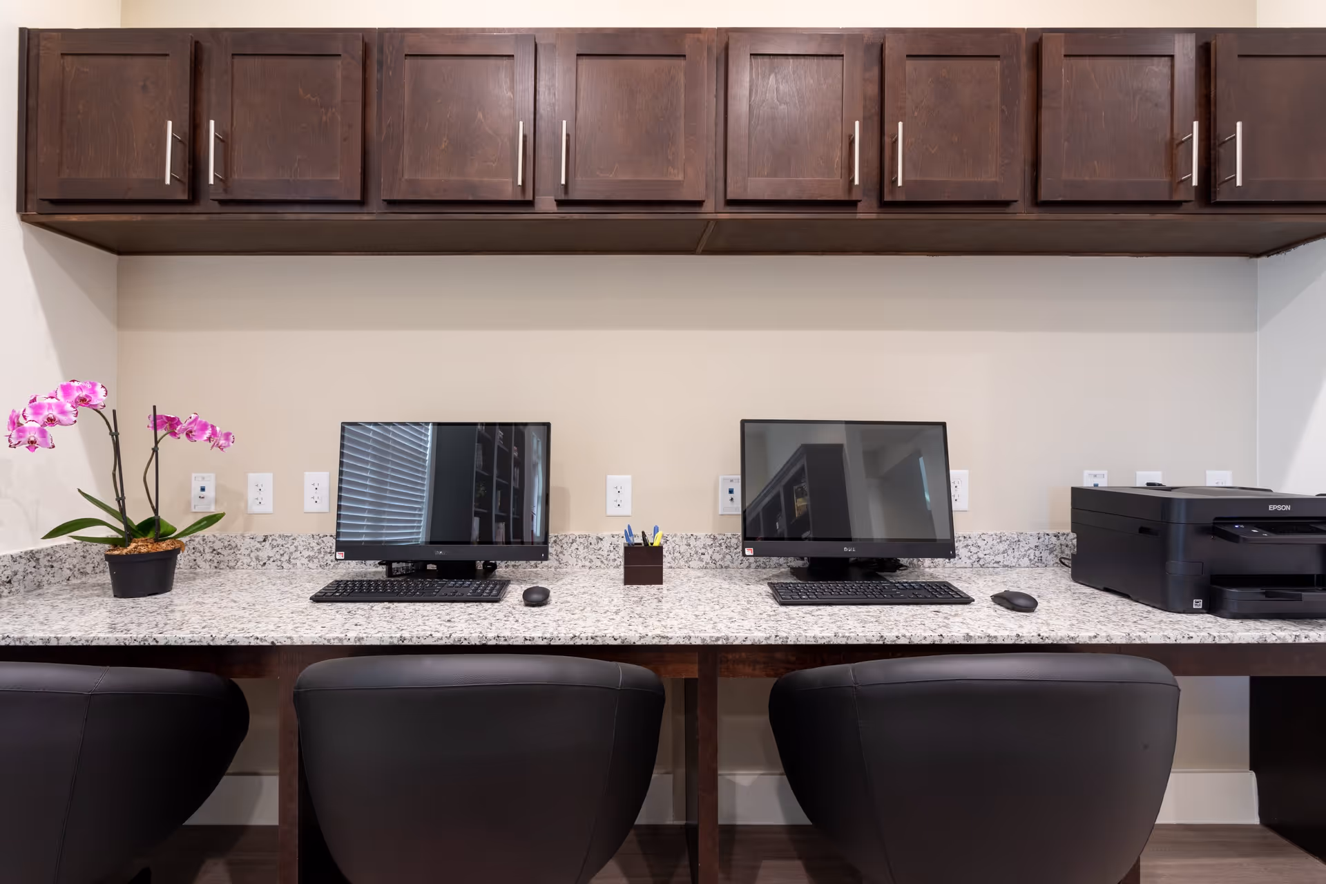 A workspace area with two computer monitors, keyboards, and mice on a granite countertop. There is a black Epson printer on the right side and a small pot with pink orchids on the left. Above the countertop are dark wooden cabinets. Three black chairs are positioned in front of the workspace.