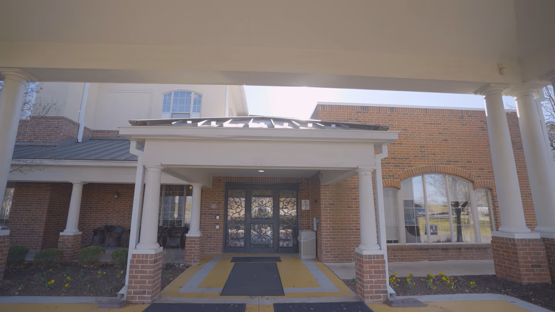 Entrance to a building with a covered porch supported by white columns. The building has red brick walls and large windows. There are small flower beds with yellow flowers on either side of the entrance.
