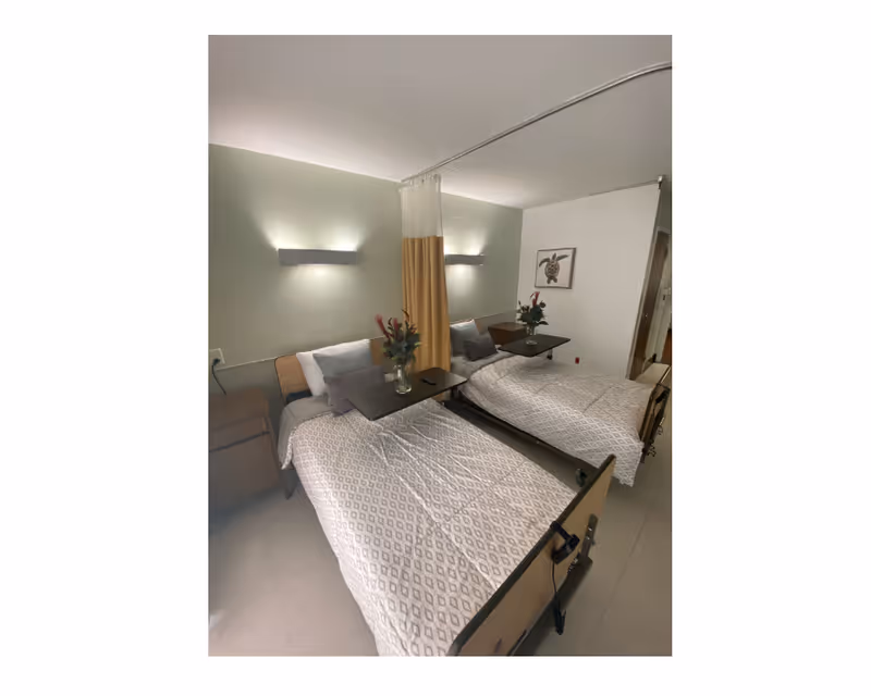 Two hospital-style beds in a shared, well-lit room with a privacy curtain and overbed tables topped with flower vases.