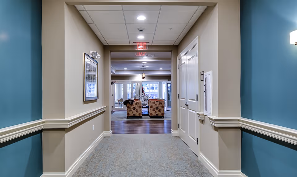 Carpeted corridor with teal accent walls and white trim opening into a seating area with patterned armchairs and an illuminated exit sign.