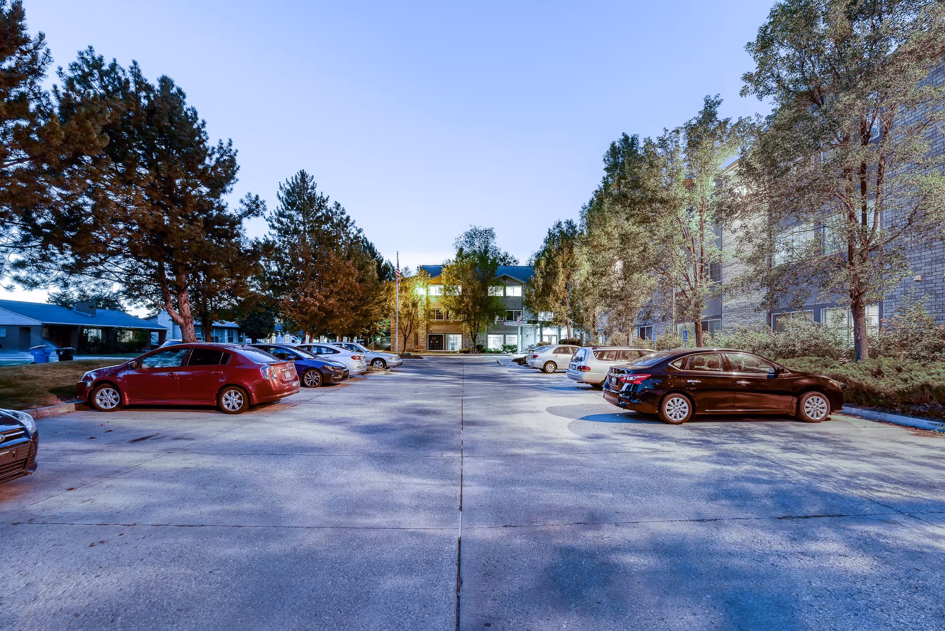 Parking lot with several parked cars on both sides, surrounded by trees and buildings, under a clear evening sky with lights illuminating the area.