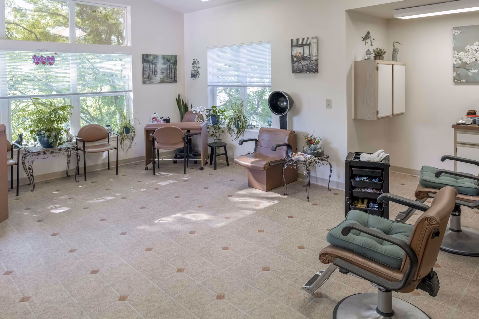 Sunlit salon-style common room with styling chairs, waiting chairs, plants and large windows.