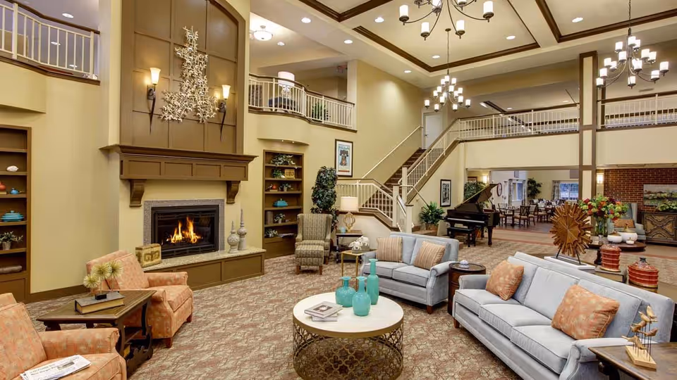 Spacious two-story communal living room with sofas and armchairs arranged around a fireplace, a grand piano, chandeliers, and an upper balcony.