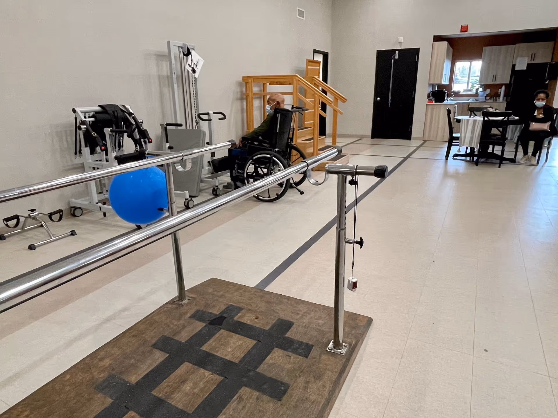 Interior view of a rehabilitation center room with parallel bars for physical therapy in the foreground. A person in a wheelchair is seated near exercise equipment along the wall. In the background, there is a small dining area with a table and chairs, and a kitchenette with cabinets and a window. Two people wearing masks are present in the room.